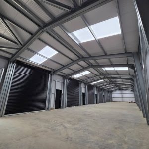 Interior view of a large steel-framed agricultural or industrial building featuring a row of black commercial roller shutter doors.