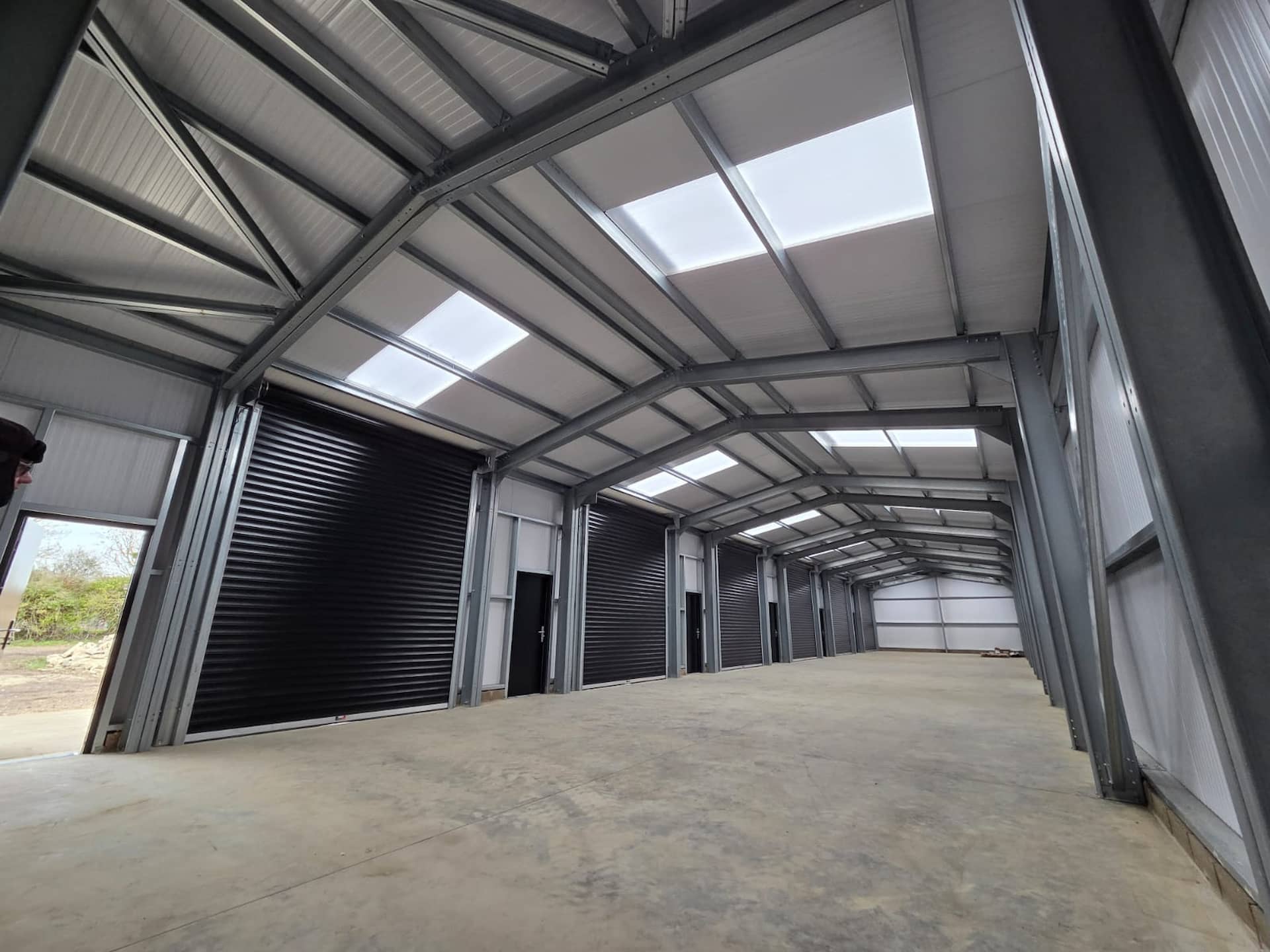 Interior view of a large steel-framed agricultural or industrial building featuring a row of black commercial roller shutter doors.