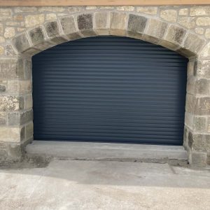 Anthracite grey insulated residential roller garage door installed seamlessly behind a traditional rustic stone archway.