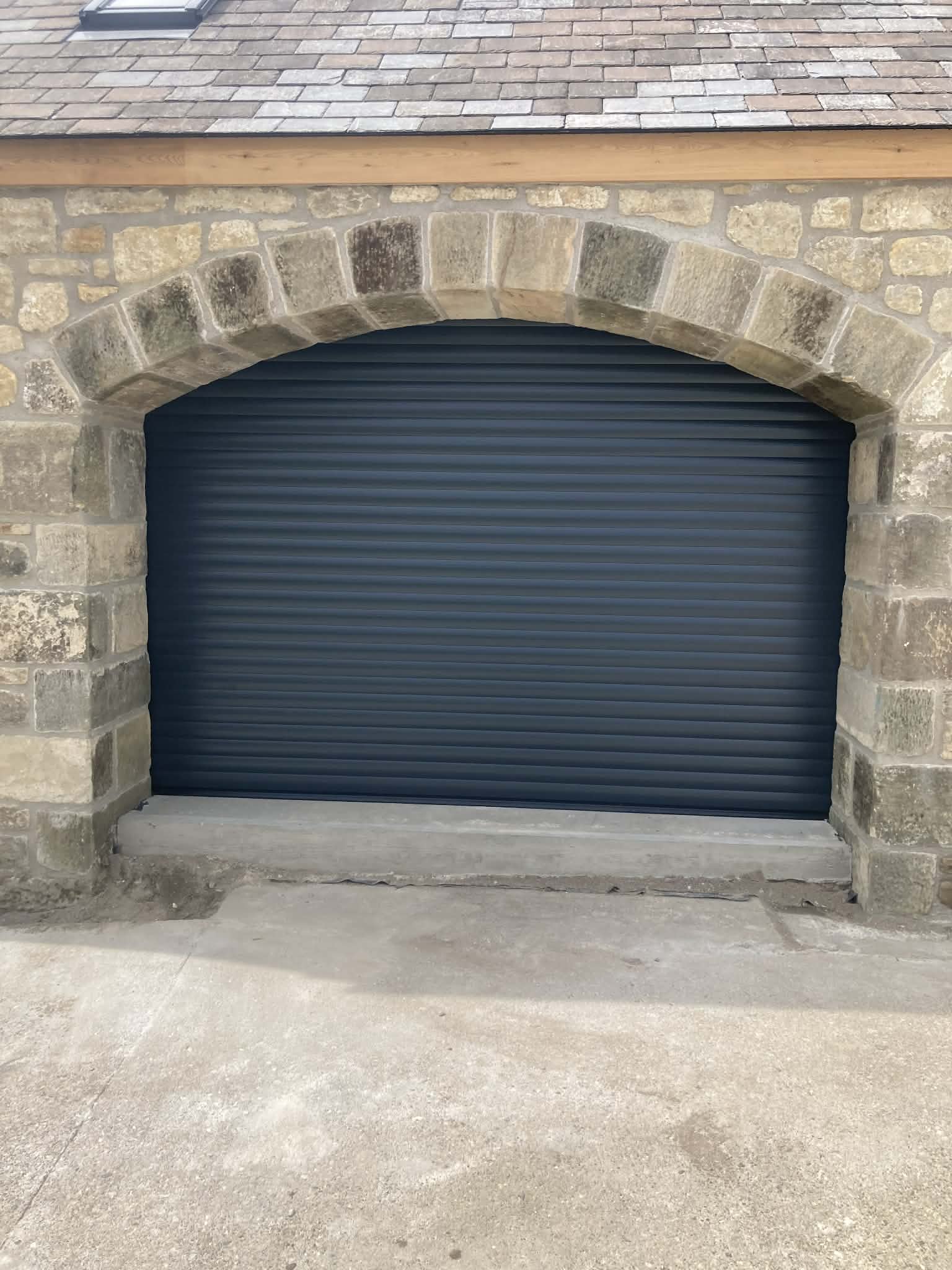 Anthracite grey insulated residential roller garage door installed seamlessly behind a traditional rustic stone archway.