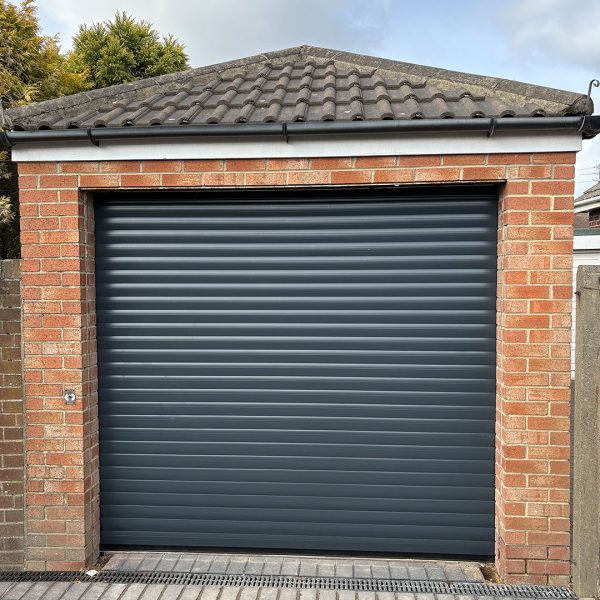 Anthracite grey residential roller garage door installed behind the brickwork of a detached brick garage with a tiled roof.