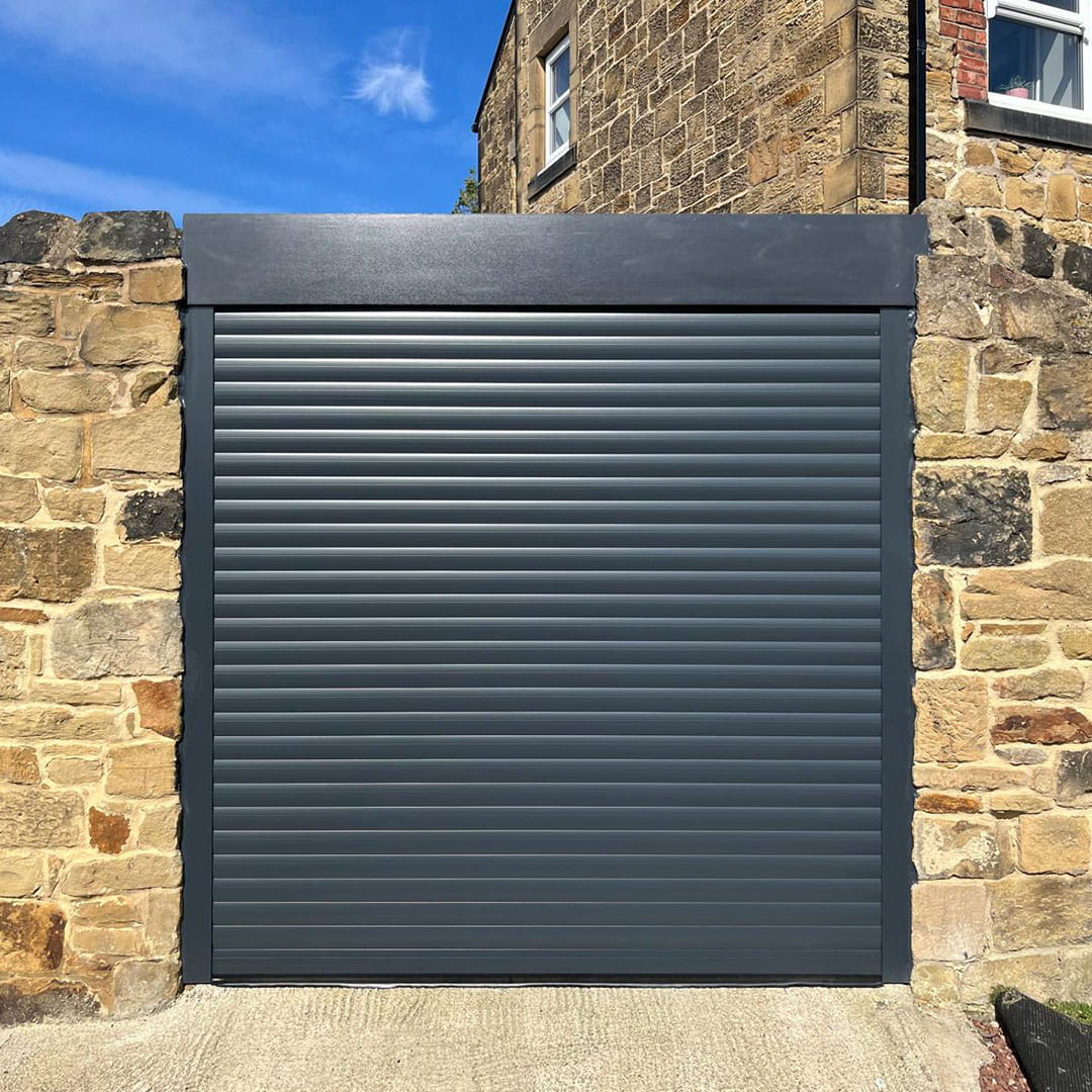 Anthracite grey insulated roller garage door with full top canopy box, installed between traditional rustic stone boundary walls.